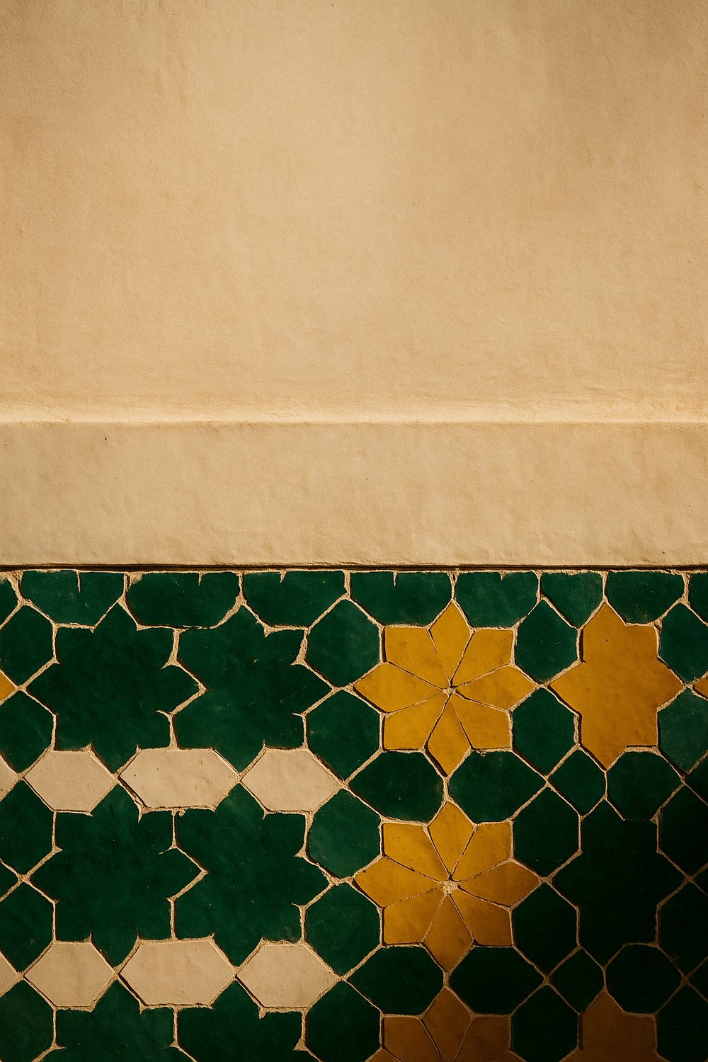 A close detail of a hand-cut zellige dado: deep glazed-green and saffron eight-point stars meeting cream tadelakt plaster above, in raking light that shows the chipped edges.